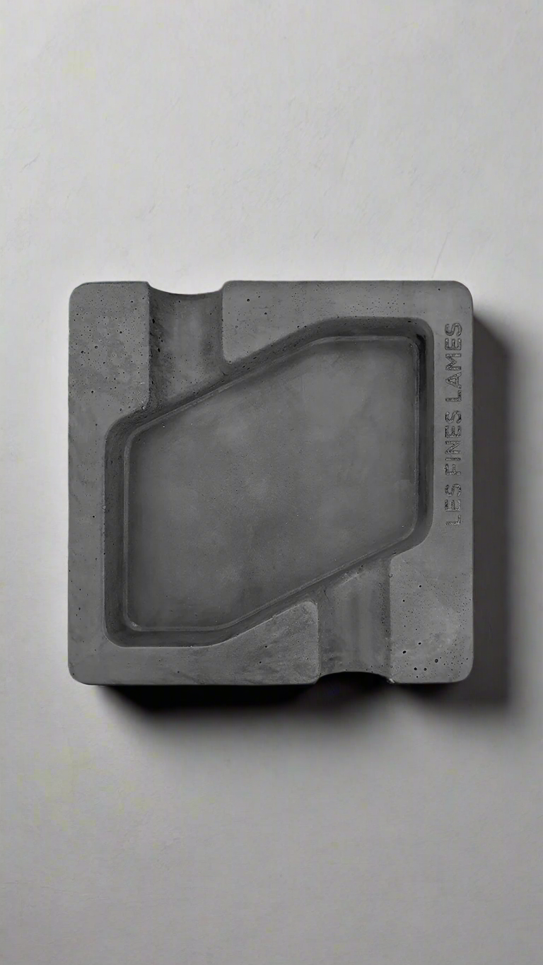 A square shaped dark grey moulded concrete ashtray with a cigar holder on opposite ends. There is a large rhombus shaped cavity in the middle of the ashtray to collect ash.