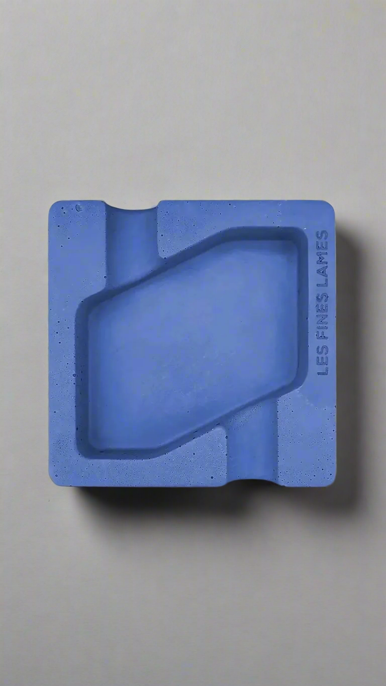 A square shaped blue moulded concrete ashtray with a cigar holder on opposite ends. There is a large rhombus shaped cavity in the middle of the ashtray to collect ash.
