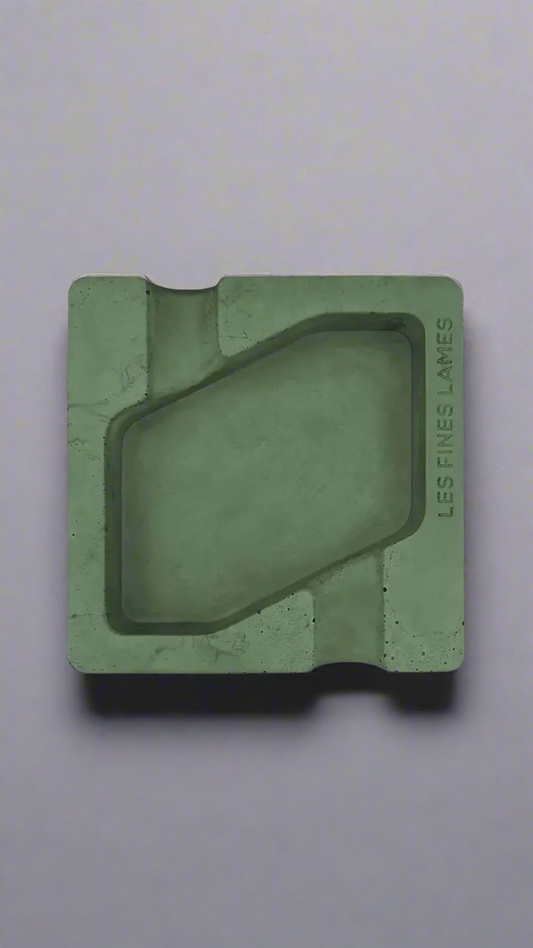 A square shaped dark green moulded concrete ashtray with a cigar holder on opposite ends. There is a large rhombus shaped cavity in the middle of the ashtray to collect ash.