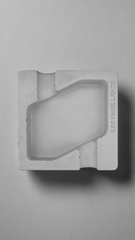 A square shaped grey moulded concrete ashtray with a cigar holders on opposite ends. There is a large rhombus shaped cavity in the middle of the ashtray to collect ash.