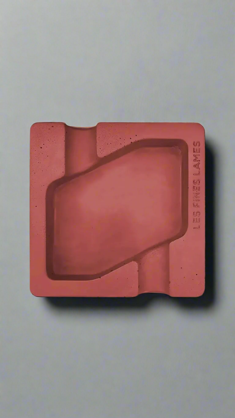 A square shaped red moulded concrete ashtray with a cigar holder on opposite ends. There is a large rhombus shaped cavity in the middle of the ashtray to collect ash.