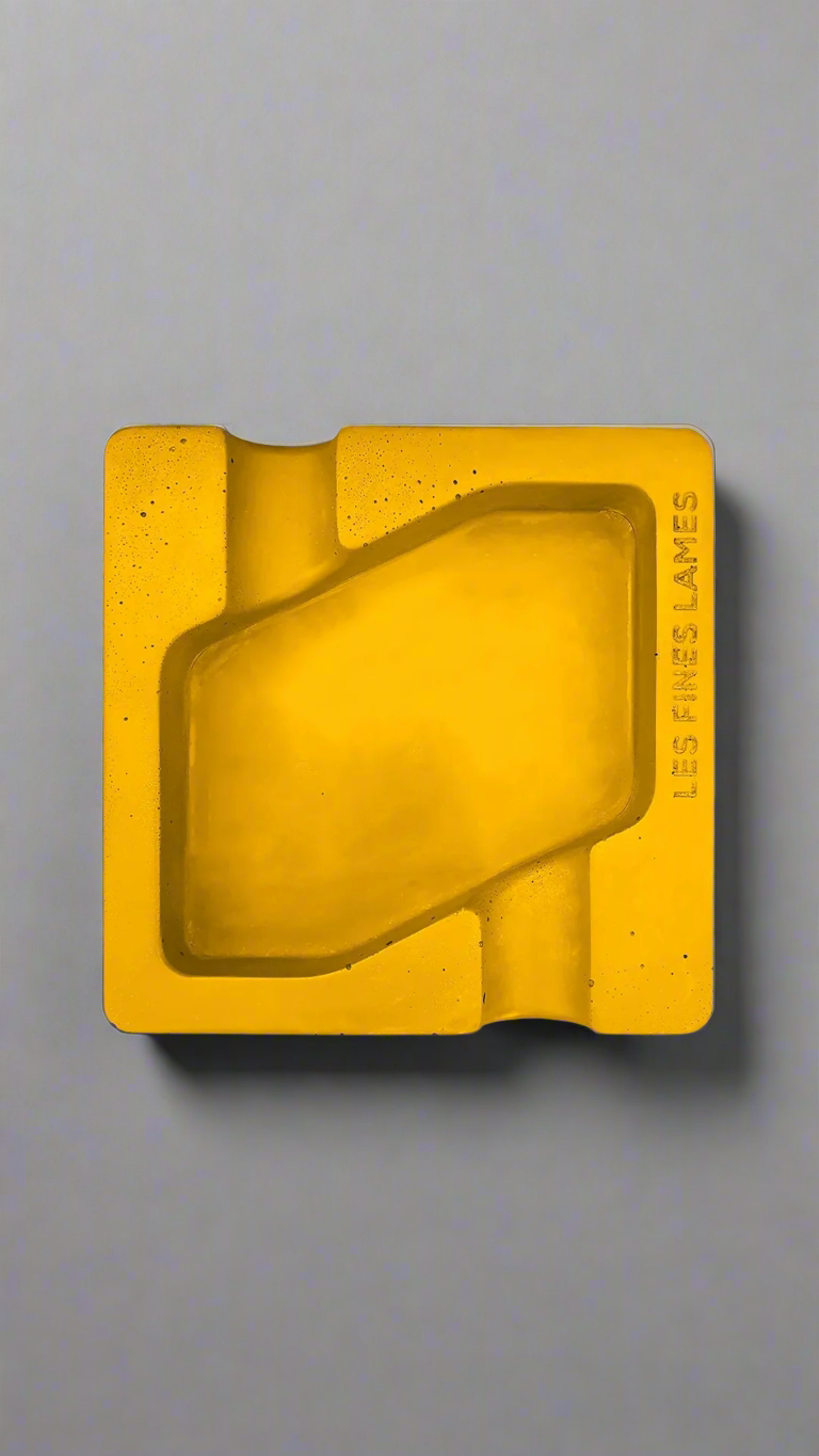 A square shaped yellow moulded concrete ashtray with a cigar holder on opposite ends. There is a large rhombus shaped cavity in the middle of the ashtray to collect ash.