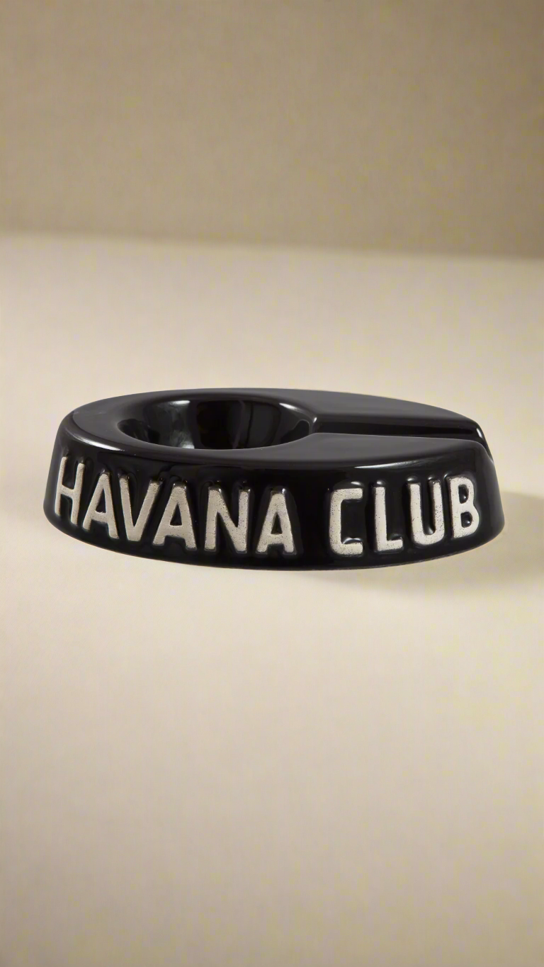 A oval shaped black glazed ceramic ashtray with a cigar holder on one of the long ends. The words Havana club are embossed on the side, the lettering is unglazed.