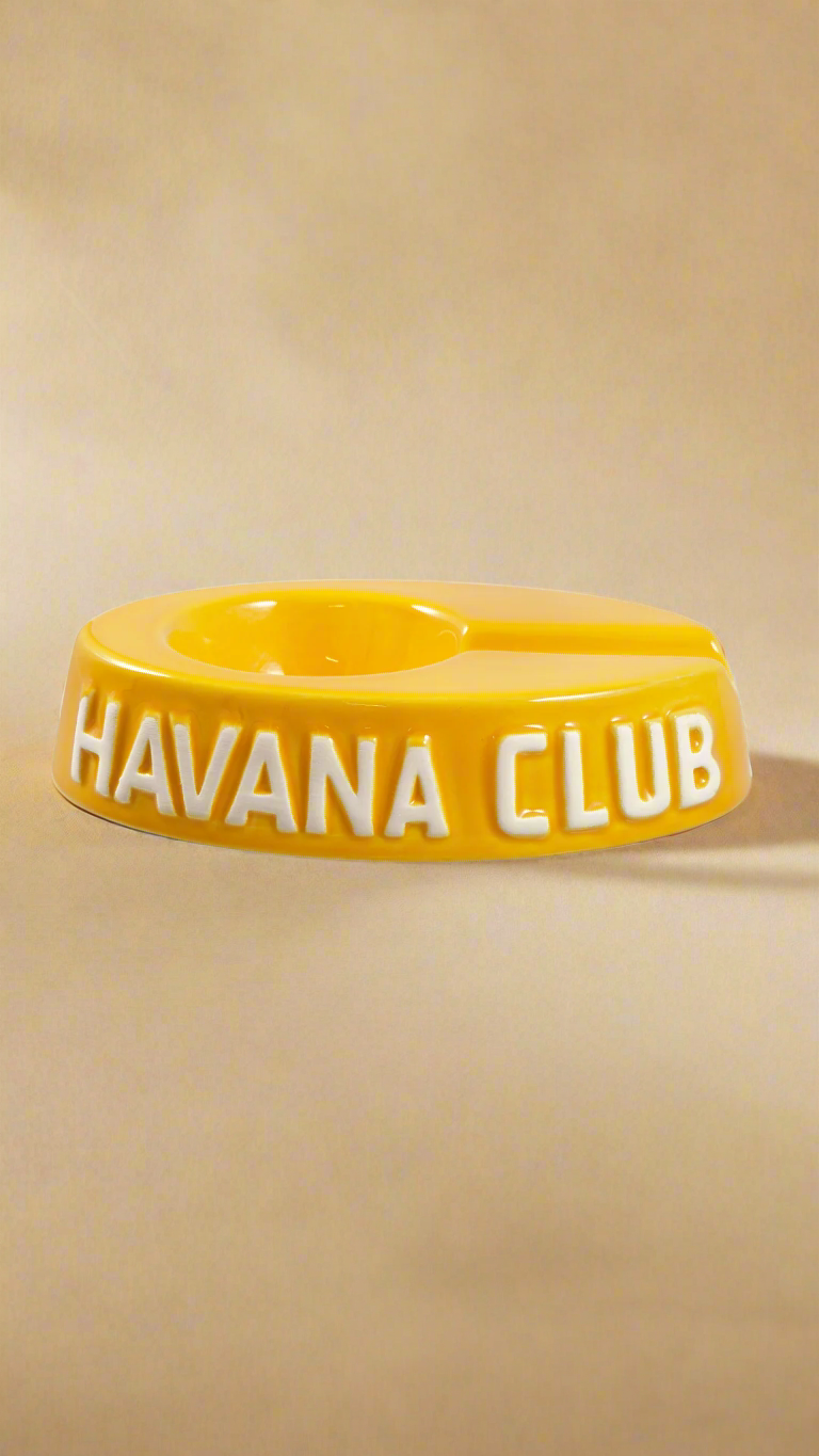 A oval shaped deep yellow glazed ceramic ashtray with a cigar holder on one of the long ends. The words Havana club are embossed on the side, the lettering is unglazed.