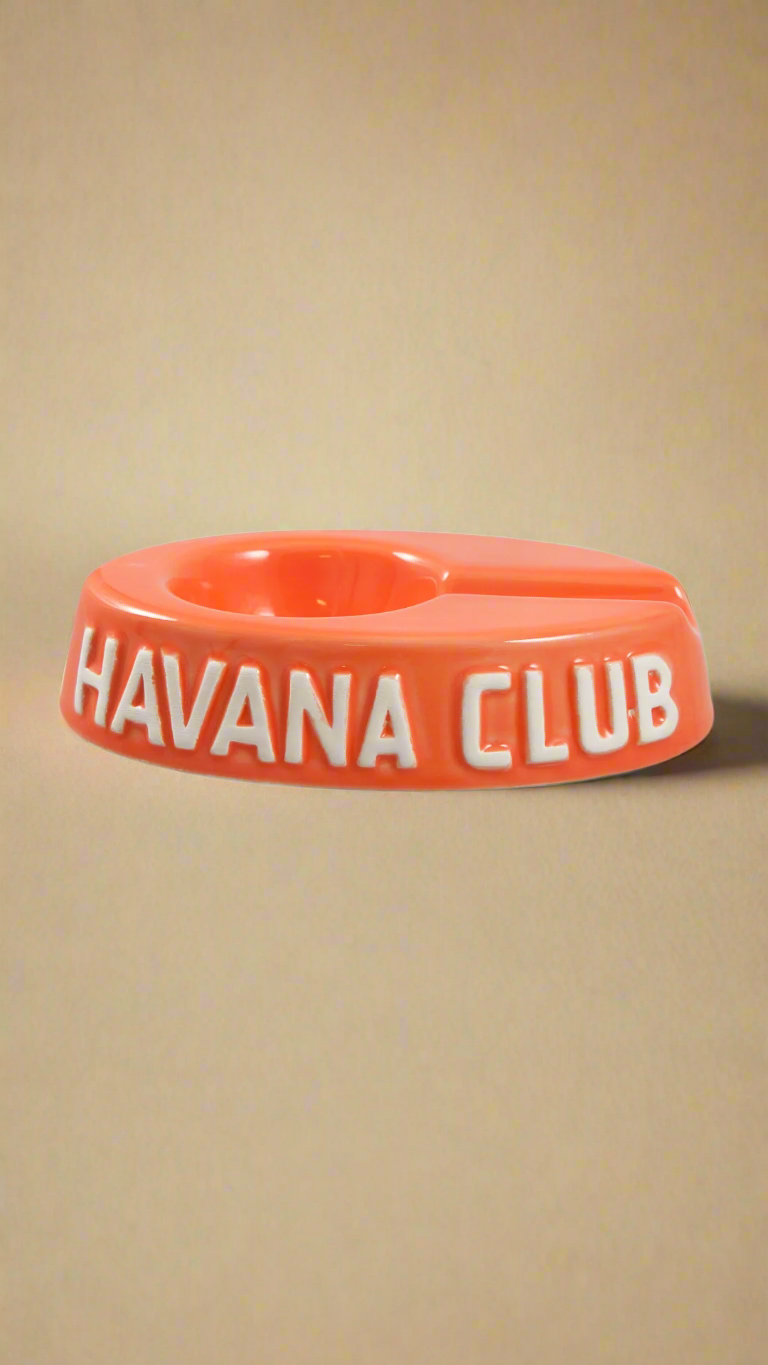 A oval shaped orange glazed ceramic ashtray with a cigar holder on one of the long ends. The words Havana club are embossed on the side, the lettering is unglazed.