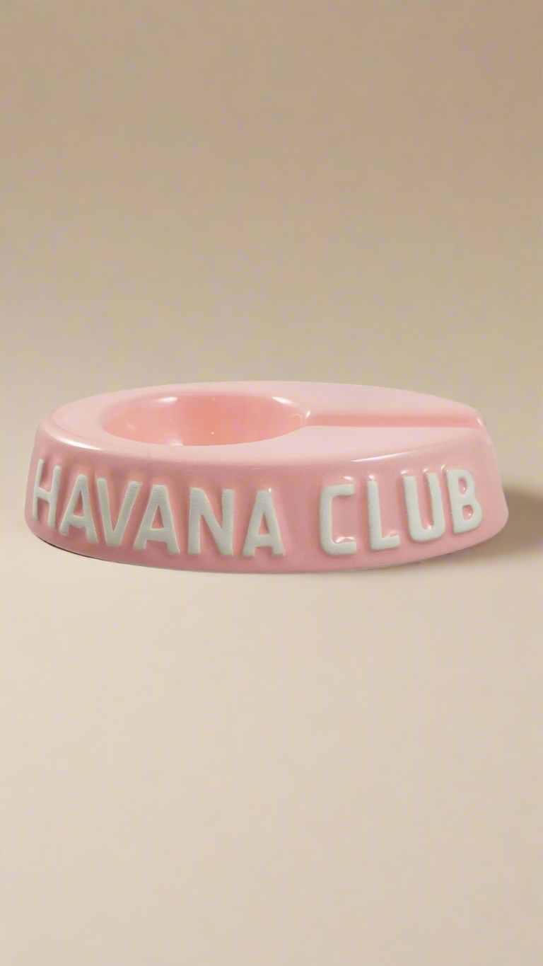 A oval shaped light pink glazed ceramic ashtray with a cigar holder on one of the long ends. The words Havana club are embossed on the side, the lettering is unglazed.