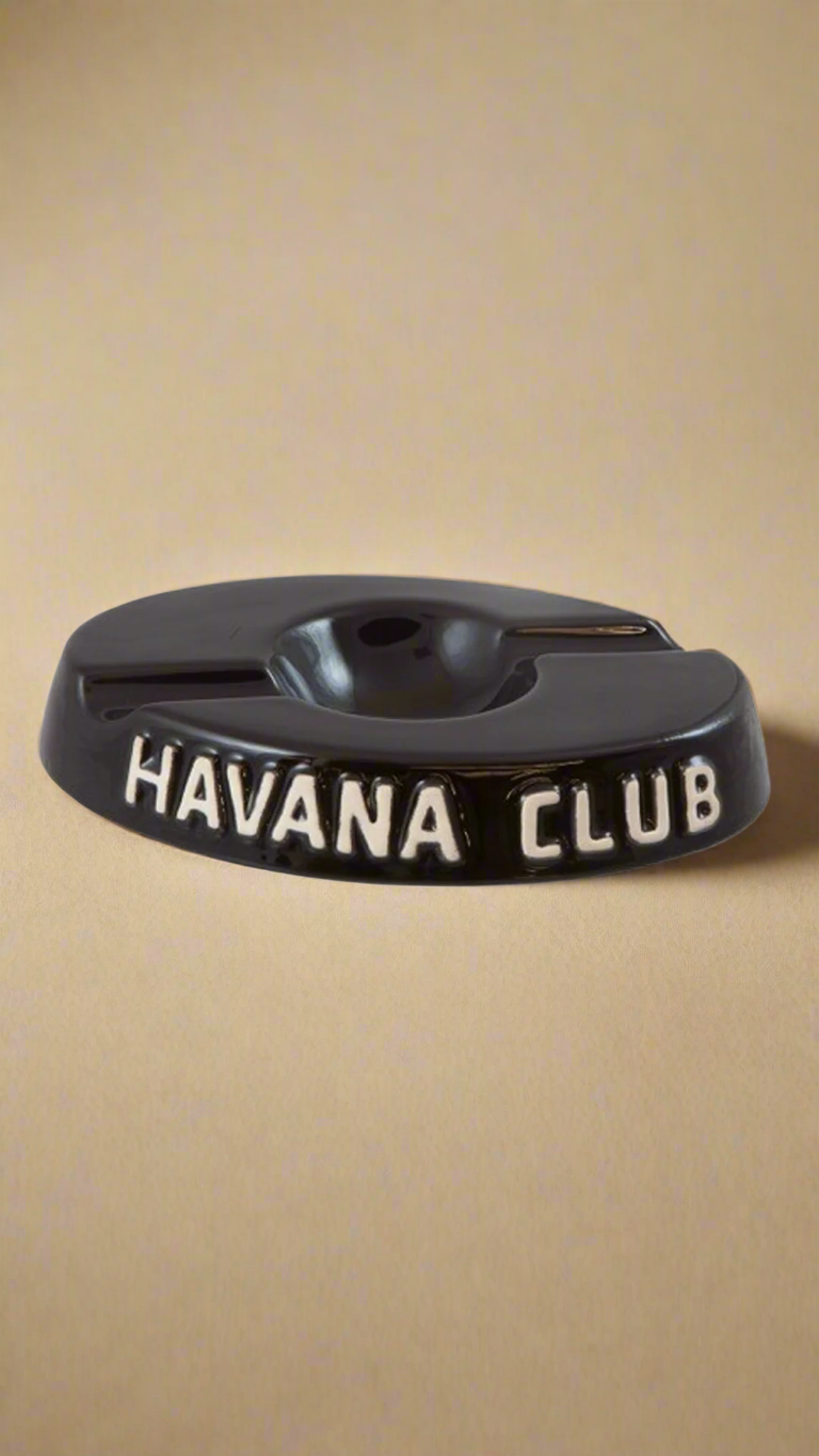 A oval shaped black glazed ceramic ashtray with two cigar holders on opposite sides. The words Havana club are embossed on the side, the lettering is unglazed.