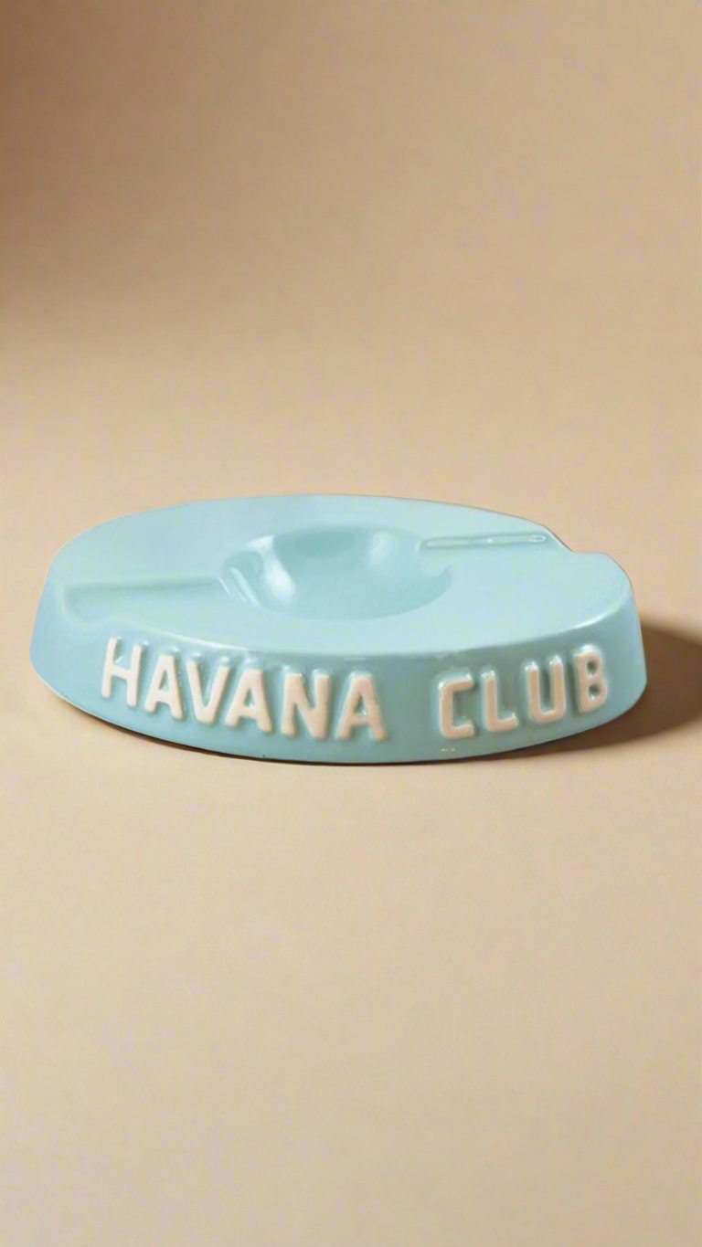 A oval shaped light blue glazed ceramic ashtray with two cigar holders on opposite sides. The words Havana club are embossed on the side, the words are in raw ceramic.