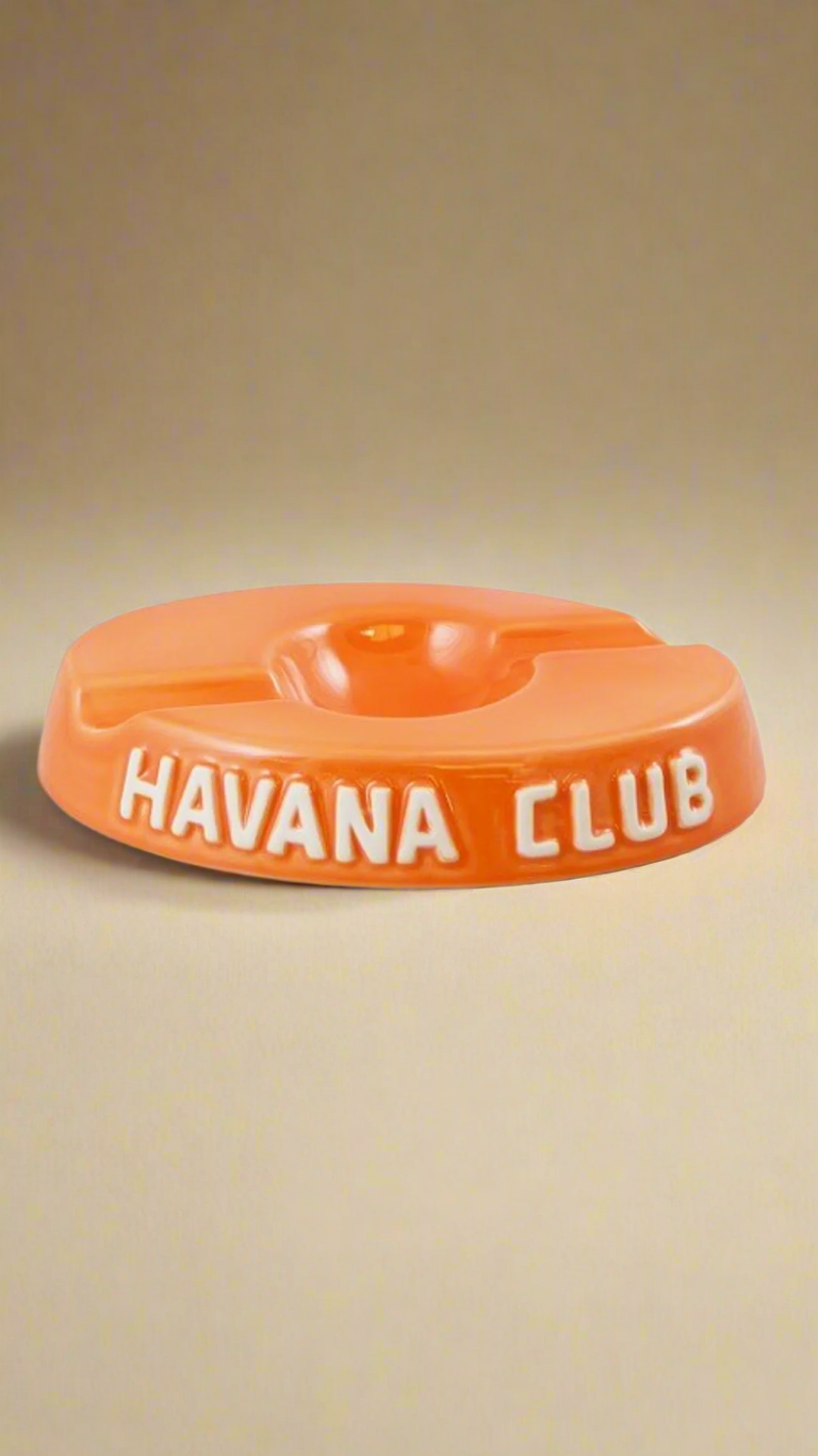 A oval shaped bright orange glazed ceramic ashtray with two cigar holders on opposite sides. The words Havana club are embossed on the side, the words are in raw ceramic.