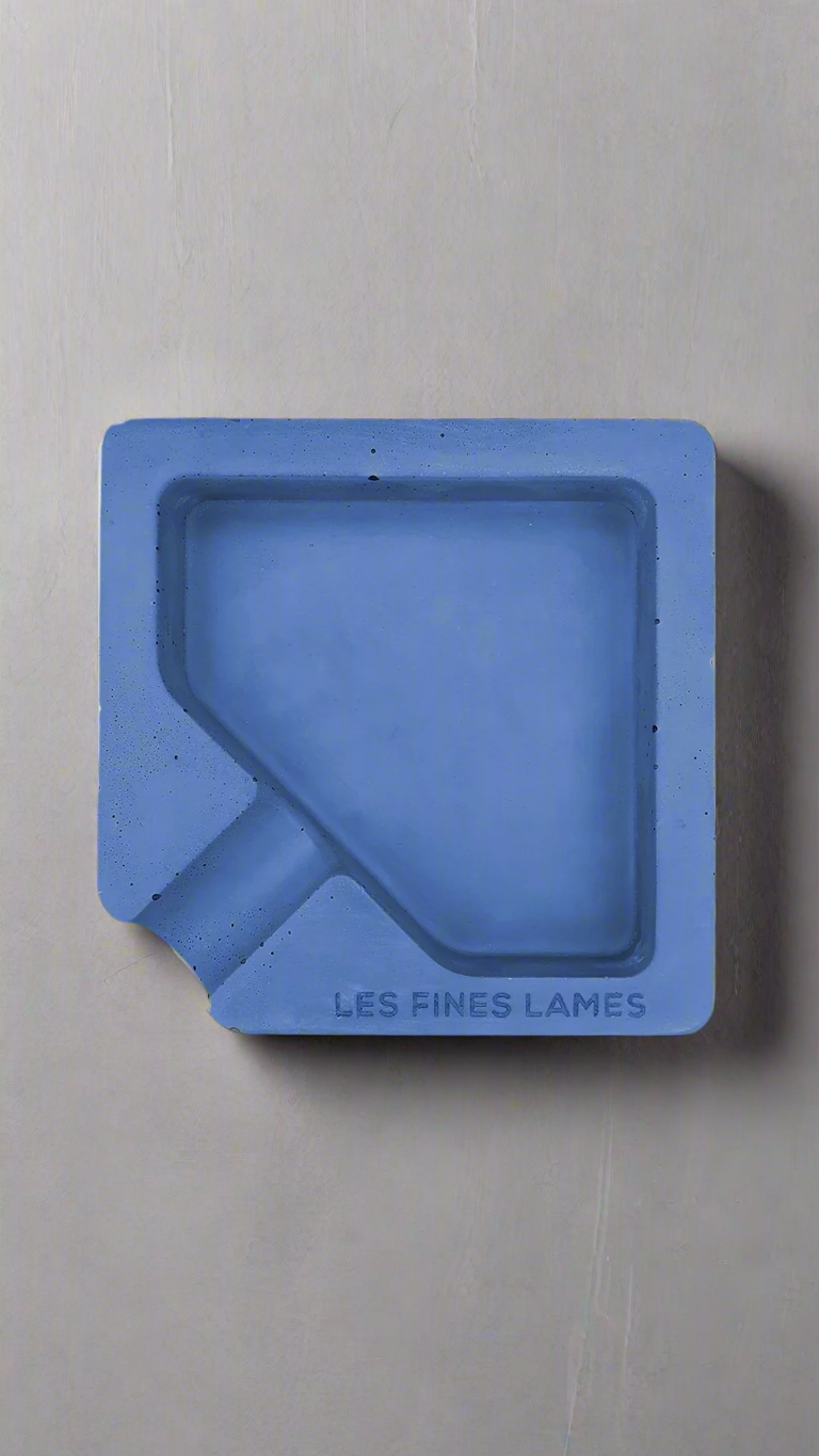 A square shaped blue moulded concrete ashtray with a cigar holder on one corner. There is a large diamond shaped cavity in the middle of the ashtray to collect ash.