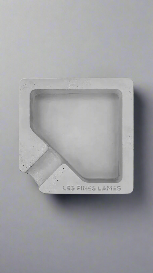 A square shaped grey moulded concrete ashtray with a cigar holder on one corner. There is a large diamond shaped cavity in the middle of the ashtray to collect ash.