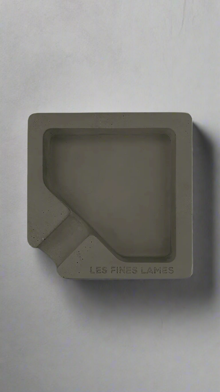 A square shaped dark green moulded concrete ashtray with a cigar holder on one corner. There is a large diamond shaped cavity in the middle of the ashtray to collect ash.