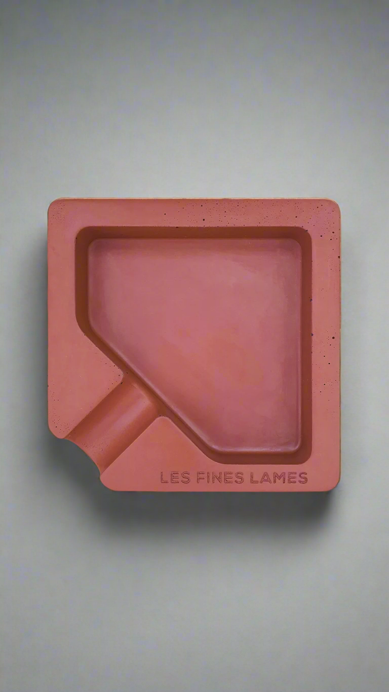 A square shaped red moulded concrete ashtray with a cigar holder on one corner. There is a large diamond shaped cavity in the middle of the ashtray to collect ash.