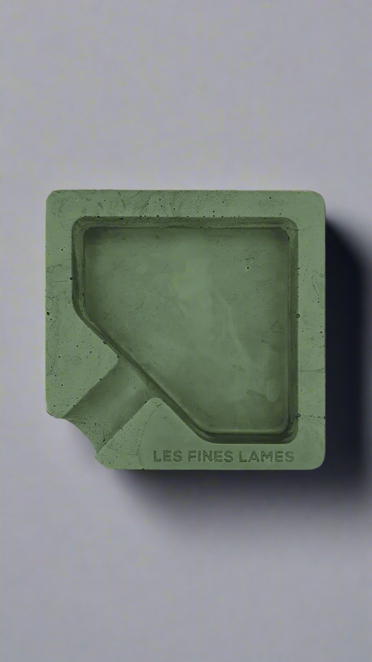 A square shaped green moulded concrete ashtray with a cigar holder on one corner. There is a large diamond shaped cavity in the middle of the ashtray to collect ash.