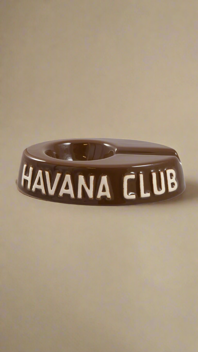 A oval shaped brown glazed ceramic ashtray with a cigar holder on one of the long ends. The words Havana club are embossed on the side, the lettering is unglazed.