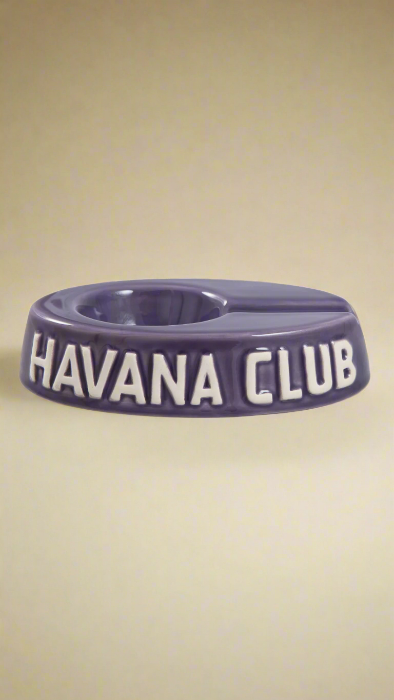 A oval shaped purple glazed ceramic ashtray with a cigar holder on one of the long ends. The words Havana club are embossed on the side, the lettering is unglazed.