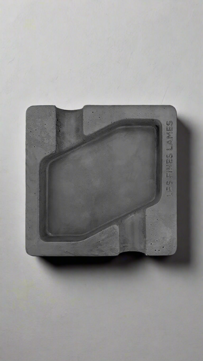 A square shaped dark grey moulded concrete ashtray with a cigar holder on opposite ends. There is a large rhombus shaped cavity in the middle of the ashtray to collect ash.