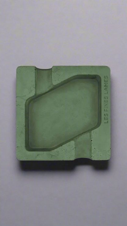 A square shaped dark green moulded concrete ashtray with a cigar holder on opposite ends. There is a large rhombus shaped cavity in the middle of the ashtray to collect ash.