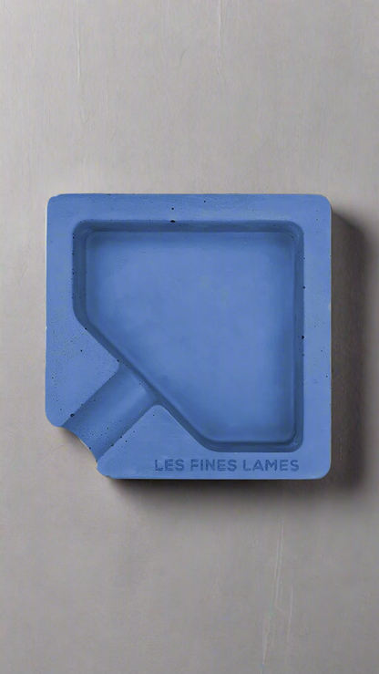 A square shaped blue moulded concrete ashtray with a cigar holder on one corner. There is a large diamond shaped cavity in the middle of the ashtray to collect ash.