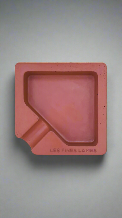 A square shaped red moulded concrete ashtray with a cigar holder on one corner. There is a large diamond shaped cavity in the middle of the ashtray to collect ash.