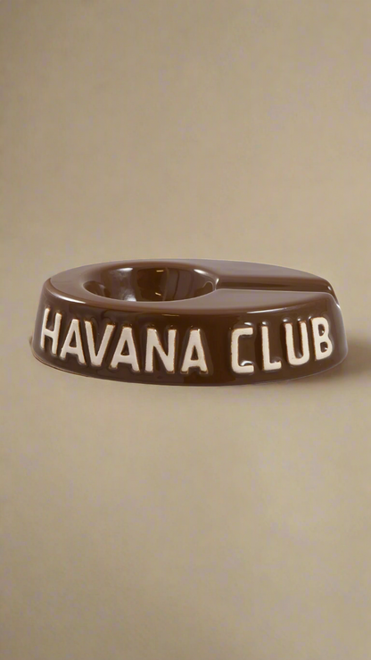 A oval shaped brown glazed ceramic ashtray with a cigar holder on one of the long ends. The words Havana club are embossed on the side, the lettering is unglazed.